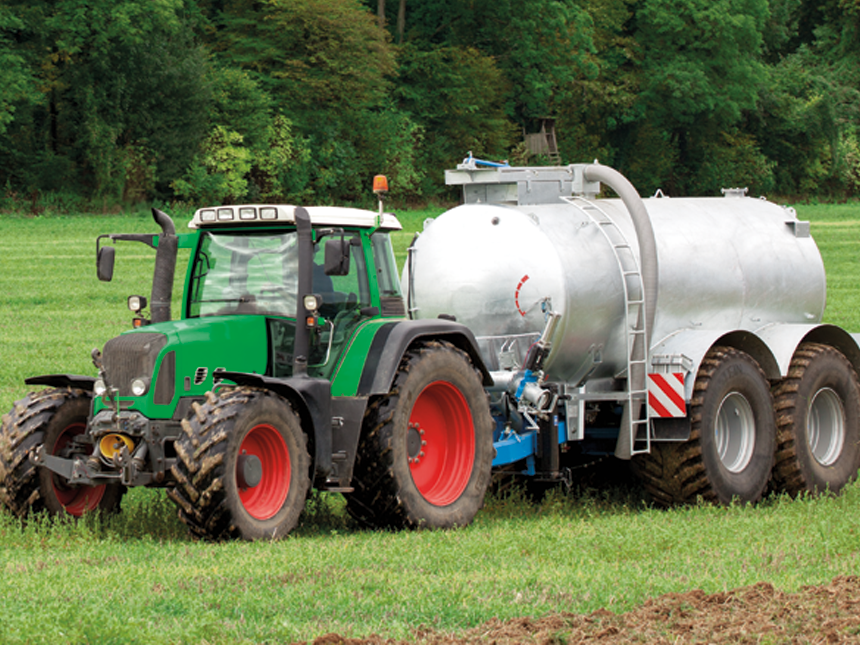 Ein Traktor zieht einen grauen Anhänger mit Güllekompressor auf einem grünen Ackerfeld