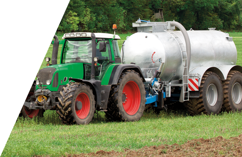 Ein Traktor zieht einen grauen Anhänger mit Güllekompressor auf einem grünen Ackerfeld