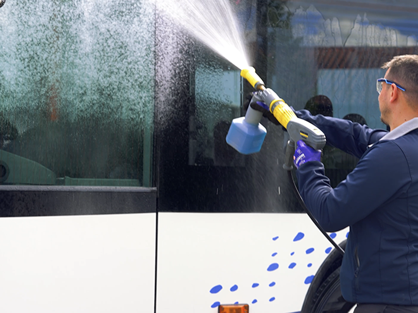 Une personne nettoie le côté d'un véhicule avec un appareil haute pression. La personne porte des gants et tient le nettoyeur haute pression à deux mains, le jet d'eau étant dirigé vers la surface du véhicule. Le véhicule semble être blanc avec des taches bleues près du sol.