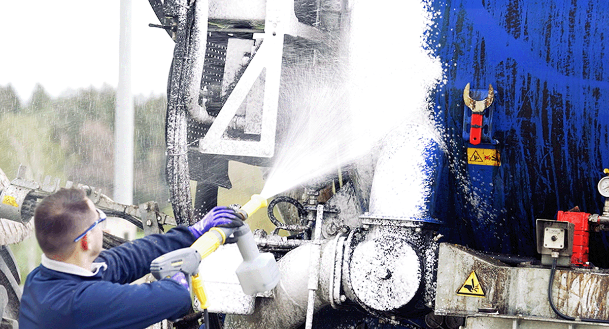 Eine Person reinigt Industriemaschinen mit einem Hochdruckreiniger. Die Maschinen scheinen mit Bau- oder Fertigungsprozessen verbunden zu sein. Die Person trägt Schutzkleidung, einschließlich Handschuhen und Schutzbrille. Auf den Maschinen sind verschiedene Komponenten und Warnschilder sichtbar.