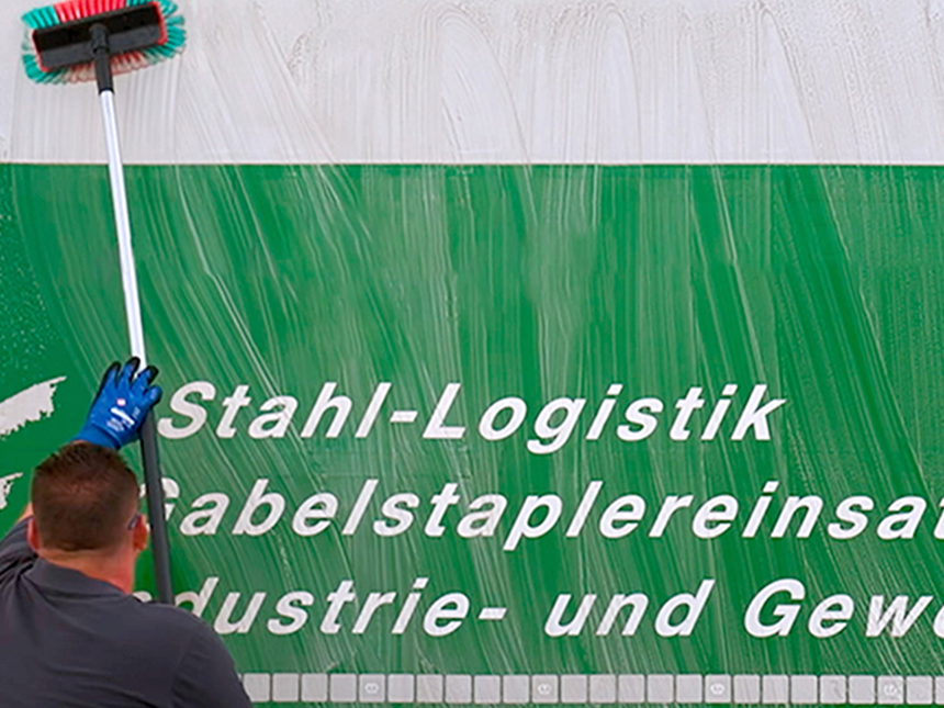 Eine Person reinigt eine grün-weiße Oberfläche mit einem langen Bürstenstiel. Die Person trägt blaue Handschuhe und ein dunkles Shirt. Auf der Oberfläche steht der Text: "Stahl-Logistik Gabelstaplereinsatz Industrie- und Gewerbe".