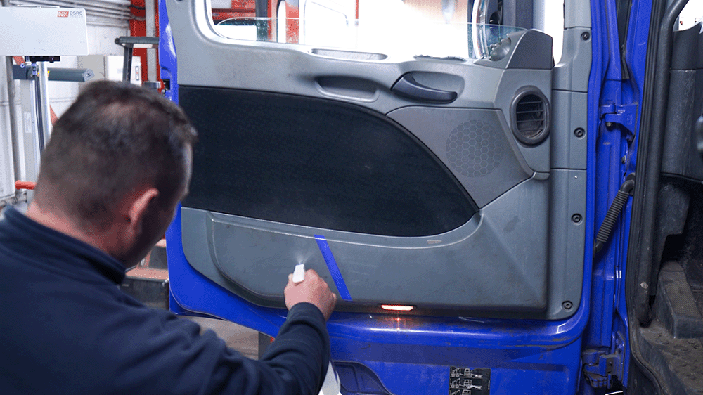 Eine Person sprüht einen Spezialreiniger auf die verschmutzte Türverkleidung eines LKW, um Schuhstreifen und Gummiabrieb zu entfernen – der betroffene Bereich wurde zuvor mit blauem Klebeband abgeklebt, um den Vorher-nachher-Effekt besser sichtbar zu machen.