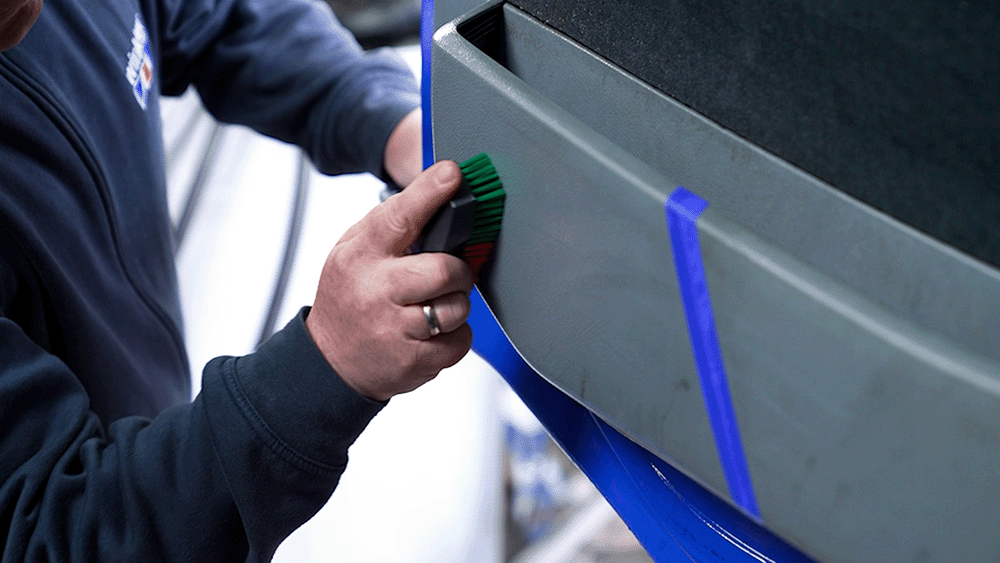 Reinigung von Schuhstreifen auf der Türverkleidung eines LKW: Eine Person entfernt mit einer Handbürste Gummiabrieb von der verschmutzten Kunststoffoberfläche – der Bereich ist durch Klebeband zur besseren Sichtbarkeit vorher und nachher abgetrennt.