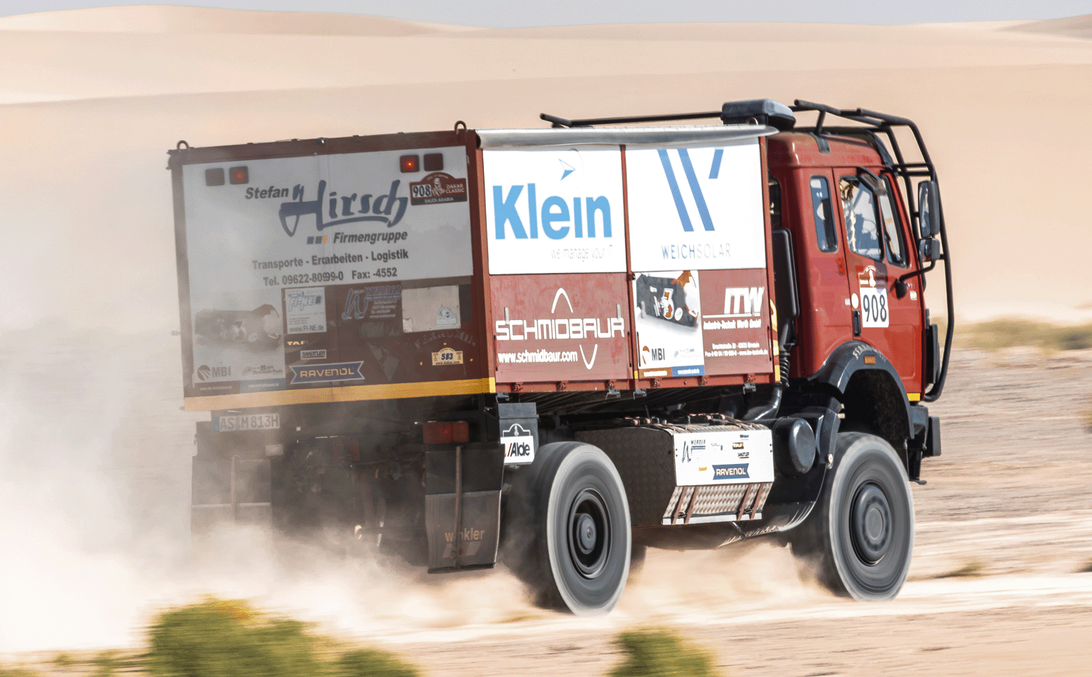 Rallye-Truck im Wüstenrennen Ein Rallye-Truck mit verschiedenen Sponsorenlogos, darunter Stefan Hirsch Firmengruppe, Klein und Schmidbauer, fährt durch eine Wüstenlandschaft.
