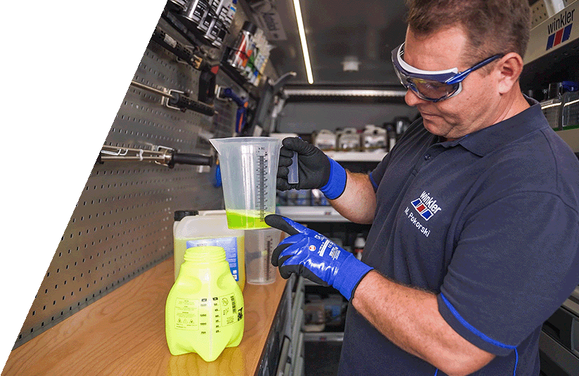 Ein winkler Mitarbeiter misst das Mischungsverhältnis eines Reinigungsmittels ab. Er trägt Schutzbrille und Handschuhe und steht in einem Servicefahrzeug mit Werkstattausrüstung.