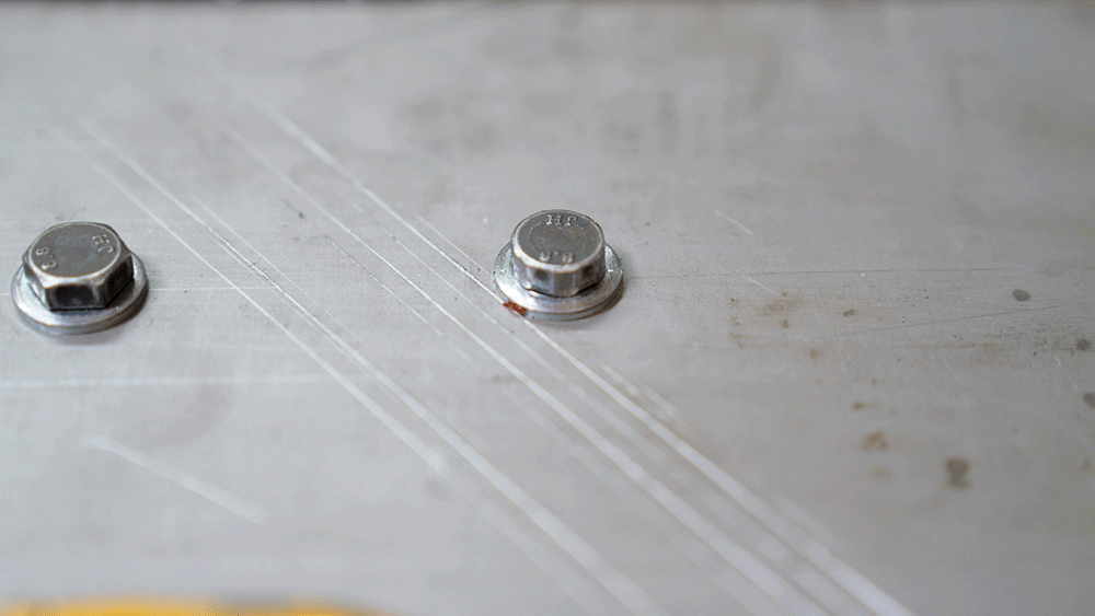Zwei Sechskantschrauben auf einer Metallplatte, eine davon mit sichtbaren Beschädigungen am Schraubenkopf.